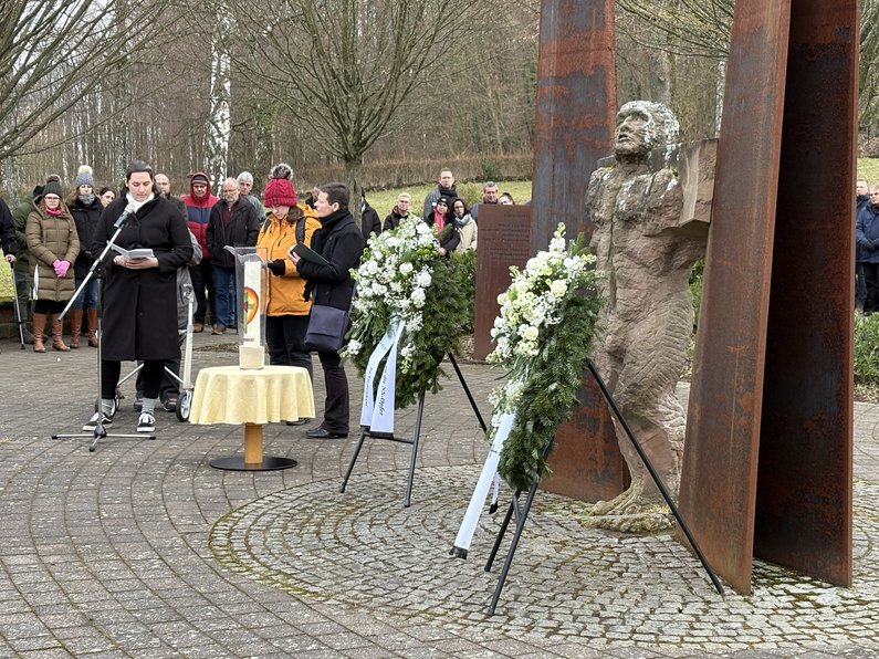 Frauen berichten in einer Andacht über das Schicksal von Maria Z. und anderen Patient*innen, im Vordergrund die Skulptur auf der Gedenkstätte mit den Kränzen, im Hintergrund Besucher*innen.
